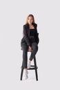 Full length portrait of beautiful young girl sitting on tall chair in black jeans and jacket over gray background Royalty Free Stock Photo