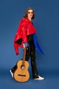 Full-length portrait of beautiful young girl in stylish clothes walking with guitar against blue studio background Royalty Free Stock Photo