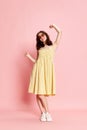 Full-length portrait of beautiful young woman in pretty yellow dress and sunglasses cheerfully dancing against pink Royalty Free Stock Photo