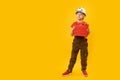 Full-length portrait of boy imitating worker. Child wears helmet with holds toolbox, isolation on yellow background. Copy space,