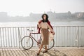 Full-length portrait of elegant woman in long skirt and blouse standing near bicycle in sunny morning. Outdoor photo of