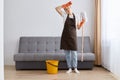 Full length portrait of exhausted dark haired woman wearing white t shirt and apron posing with mop and bucket, being tired,