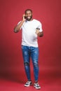 Full length portrait of handsome African American man with mobile phone and take away coffee cup. Isolated over red background Royalty Free Stock Photo