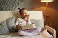 Full length portrait of hungry adorable female kid sitting on cough with crossed legs and eating fast food, holding slice of pizza Royalty Free Stock Photo