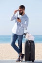 Full length man standing on beach with suitcase and mobile phone Royalty Free Stock Photo