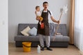 Full length portrait of man wearing casual attire and brown apron cleaning house with baby and screaming, looking at camera with