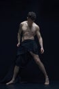 Full-length portrait of muscular, shirtless, handsome man posing in dark fabric against black studio background. Statue Royalty Free Stock Photo