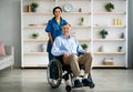 Full length portrait of older disabled man in wheelchair and his young caregiver smiling at camera in retirement home