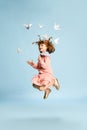 Full-length portrait of playful, lovely little girl in pink dress posing, playing with paper birds against blue studio