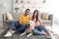 Full length portrait of positive international couple with their pet dog sitting on soft carpet at home