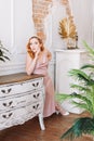 a full-length portrait of a red-haired woman in a beige silk dress leaning her elbow on an old chest of drawers in a Royalty Free Stock Photo