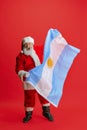 Full-length portrait of senior man wearing Santa Claus costume, holding flag of Uruguay  over red background Royalty Free Stock Photo