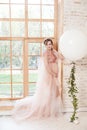 Beautiful pregnant young woman in pink dress standing near big natural window and touching her neck with hand Royalty Free Stock Photo