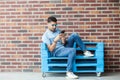 Full length portrait of serious handsome young bearded man in casual style and eyeglasses sitting on blue wooden pallet, holding Royalty Free Stock Photo