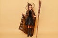 Full length portrait of serious little girl in black cloak clothing with broom isolated over beige background mysterious sorceress