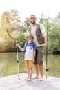 Full length portrait of smiling father and son standing with fishing tackles on pier against lake Royalty Free Stock Photo