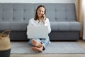 Full length portrait of smiling woman wearing white shirt and jeans sitting on floor near sofa, using laptop and talking phone, Royalty Free Stock Photo