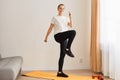 Full length portrait of woman wearing white t shirt and black leggins doing sport exercises at home, standing on mat and matching Royalty Free Stock Photo