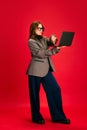 Full length portrait of young elegance woman, student in glasses working online in laptop against red studio background. Royalty Free Stock Photo