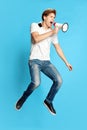 Full-length portrait of young man in casual clothes jumping and emotionally shouting in megaphone against blue studio Royalty Free Stock Photo