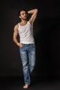 Full-length portrait of young muscular man in white undershirt and jeans posing isolated on dark background. Royalty Free Stock Photo