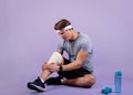 Full length portrait of young sportsman having knee injury, holding his sprained bandaged leg, violet studio background Royalty Free Stock Photo