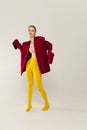 Full-length portrait of young stylish girl in yellow tights and red jacket posing isolated over grey studio background Royalty Free Stock Photo