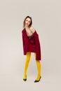 Full-length portrait of young stylish girl in yellow tights and red jacket posing isolated over grey studio background Royalty Free Stock Photo