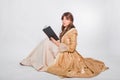 Full length portrait of a young woman in a puffy gold dress in the rococo era, posing while sitting with a book in her hands Royalty Free Stock Photo