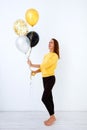 Full-length portrait of a young woman in a yellow sweater holding a bunch of balloons in gold, white and silver against a white Royalty Free Stock Photo