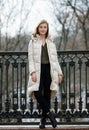 Full length posing beautiful young blonde woman standing on the spring city park in warm clothes. Cold season lifestyle freshness