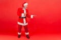 Full length positive happy man in santa claus clothes pointing finger away showing empty space for advertisement, freespace