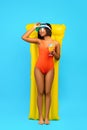 Full length of relaxed black lady in swimsuit lying on yellow inflatable lilo and drinking cocktail over blue background Royalty Free Stock Photo