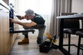 Full length shot of aged repairman in uniform working, examining broken oven in the kitchen using flashlight. Repair