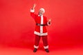 Full length shot of excited elderly man with gray beard wearing santa claus costume showing rock and roll sing to camera and Royalty Free Stock Photo