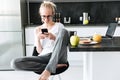 Full-length shot of young focused lady using smartphone while sitting in kitchen Royalty Free Stock Photo