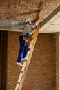 Full length shot of young workman in blue overalls and hard hat looking focused, climbing up the ladder while working on