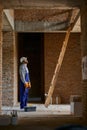 Full length shot of young workman in blue overalls and hard hat looking up while going to climb up the ladder, working