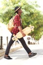 Side portrait of cool young african guy in hat walking outdoors with skateboard and bag Royalty Free Stock Photo