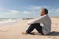 Full length side view of biracial senior man sitting on sand at beach during sunny day