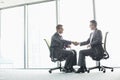 Full-length side view of businessmen shaking hands while sitting on office chairs by window Royalty Free Stock Photo