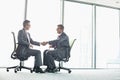 Full-length side view of businessmen shaking hands while sitting on office chairs by window Royalty Free Stock Photo