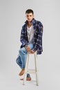 Full length studio portrait of young bold man in shirt and jeans sitting on tall wooden chair Royalty Free Stock Photo