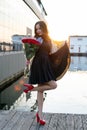 Full length view of attractive young woman with brown hair in little black dress posing and holding a bouquet of red roses at Royalty Free Stock Photo