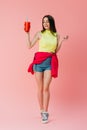 Length view of beautiful happy girl in bright clothes holding soda in plastic cup isolated on pink