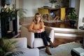 Mid aged woman using earphones and cellphone and sitting in an armchair at home Royalty Free Stock Photo