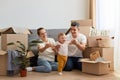 Full length of young couple moving in new apartment, sitting on floor near sofa and playing with their toddler daughter, Royalty Free Stock Photo