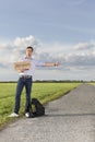 Full length of young man hitching while holding anywhere sign on countryside