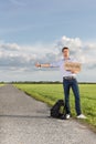 Full length of young man hitching while holding anywhere sign on countryside