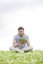 Full length of young man using digital tablet while sitting on grass against sky Royalty Free Stock Photo